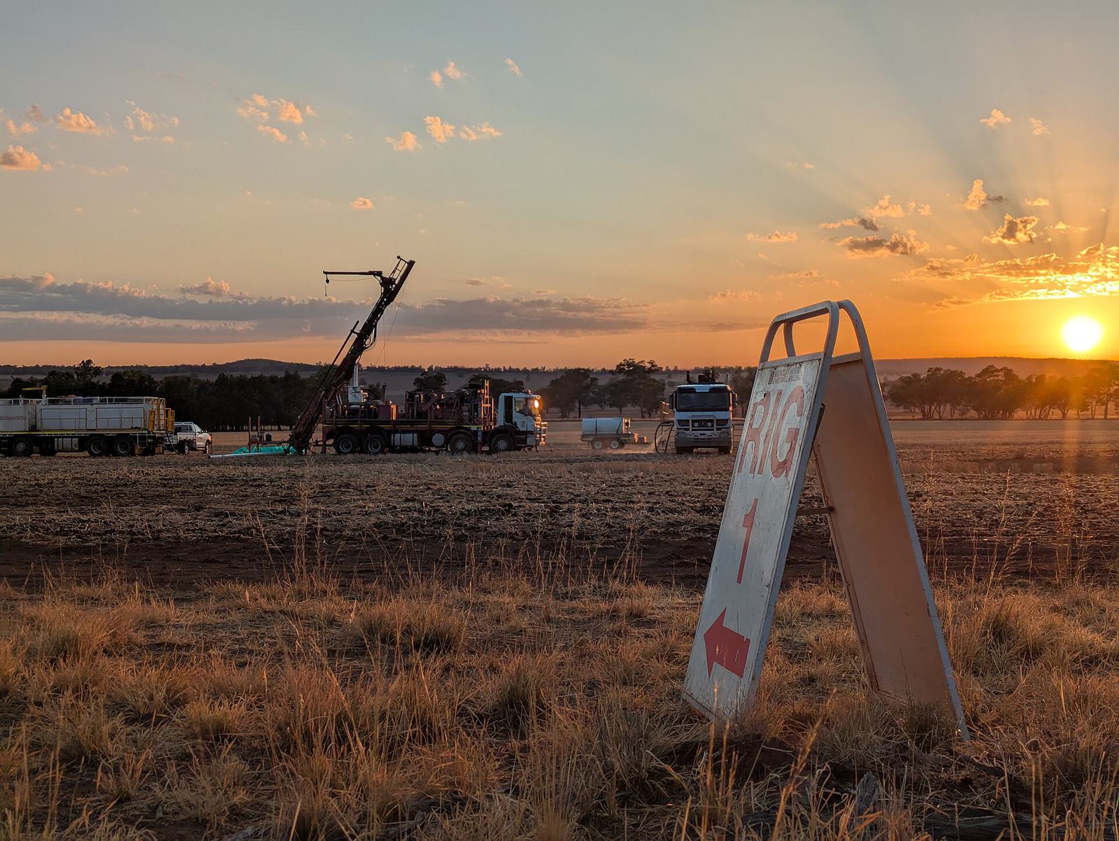 A drill rig on flat scrubland at sunrise, with a sign pointing to the site