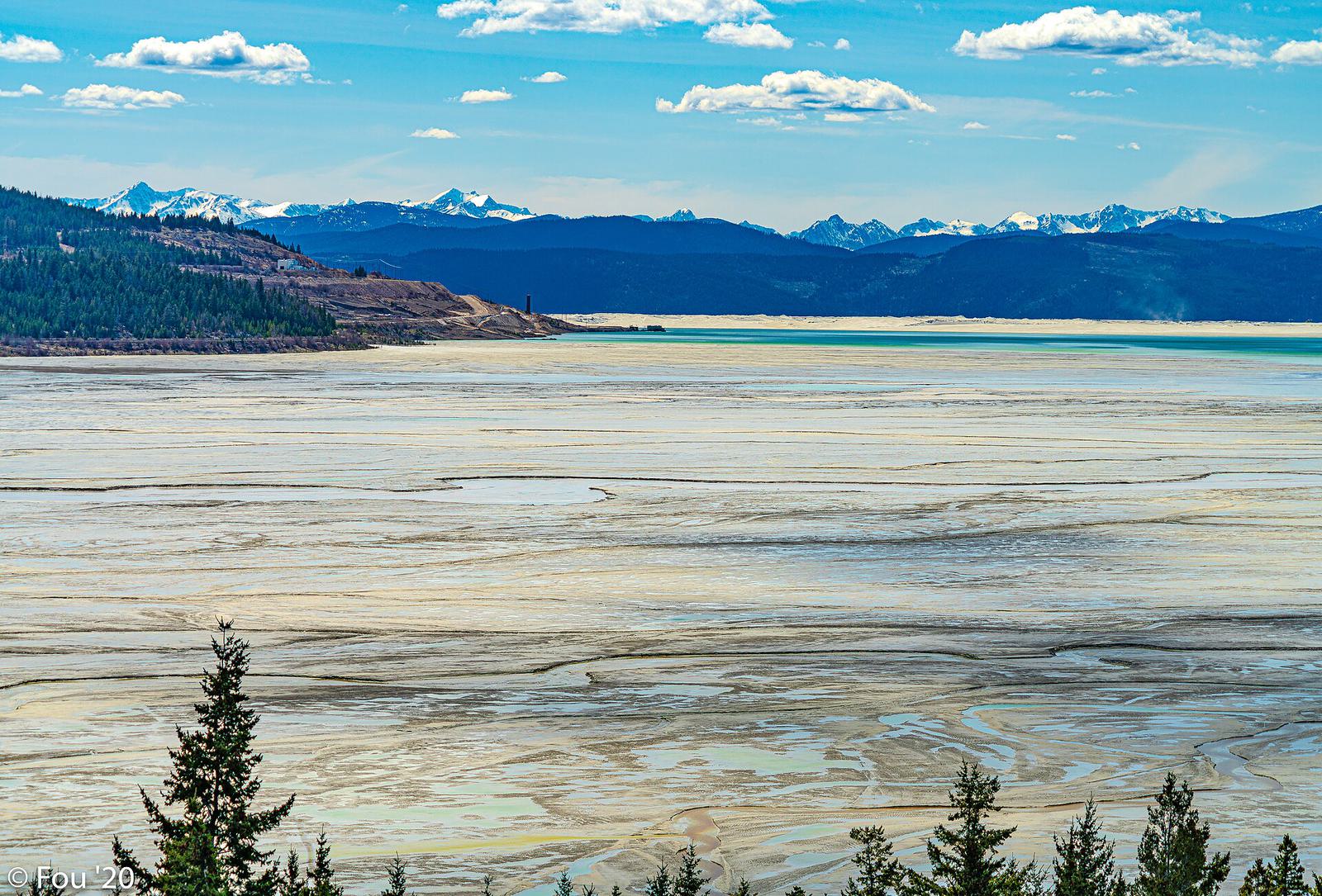 A wide pale tailings pond with forest and snowy mountains behind