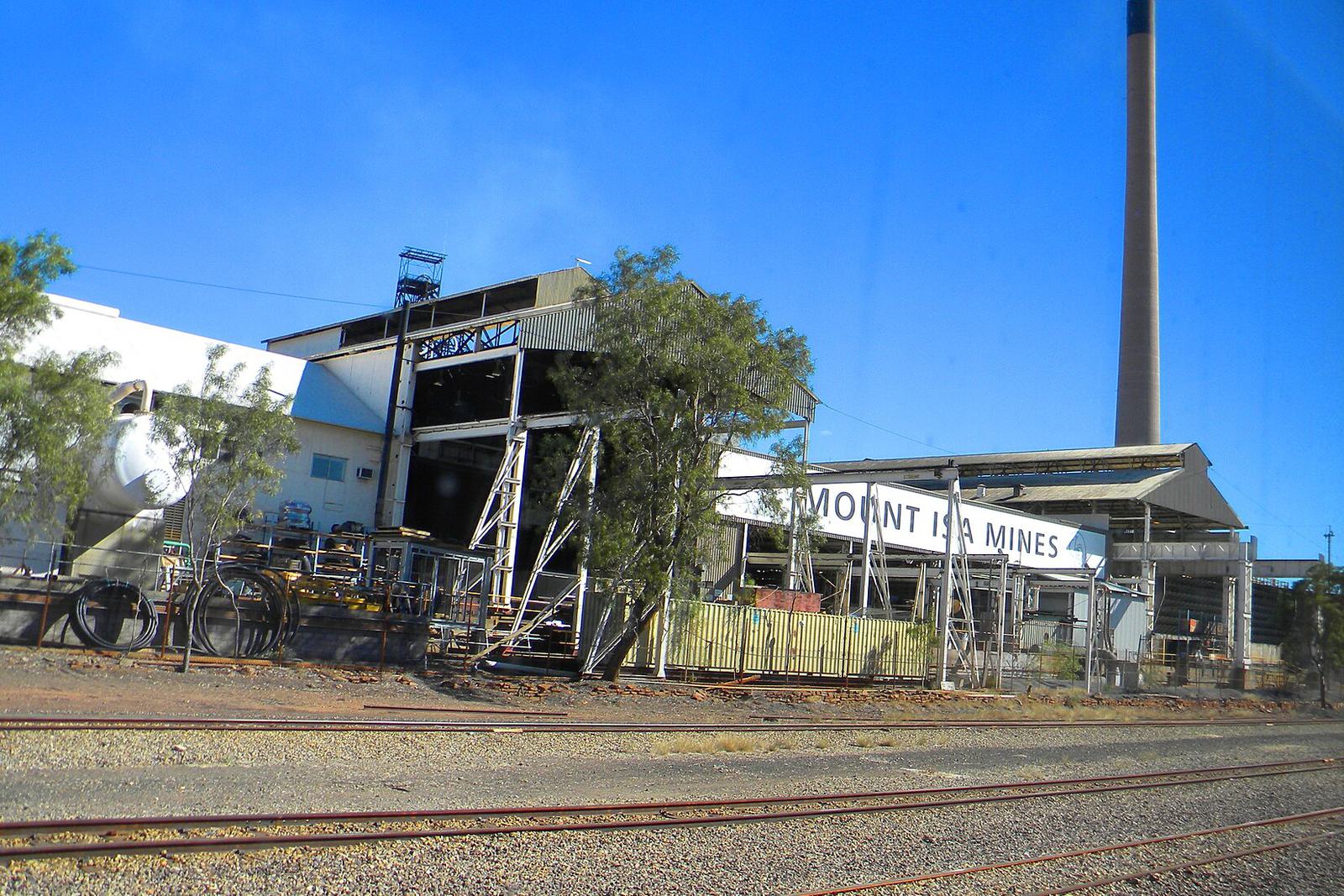 Plant buildings marked Mount Isa Mines and a tall smelter stack beside rail tracks